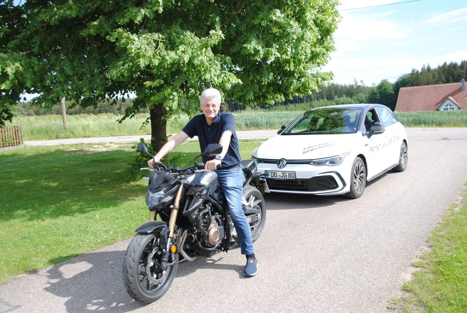 Fahrlehrer auf dem Schulungsmotorrad, dahinter das weiße Fahrschulauto