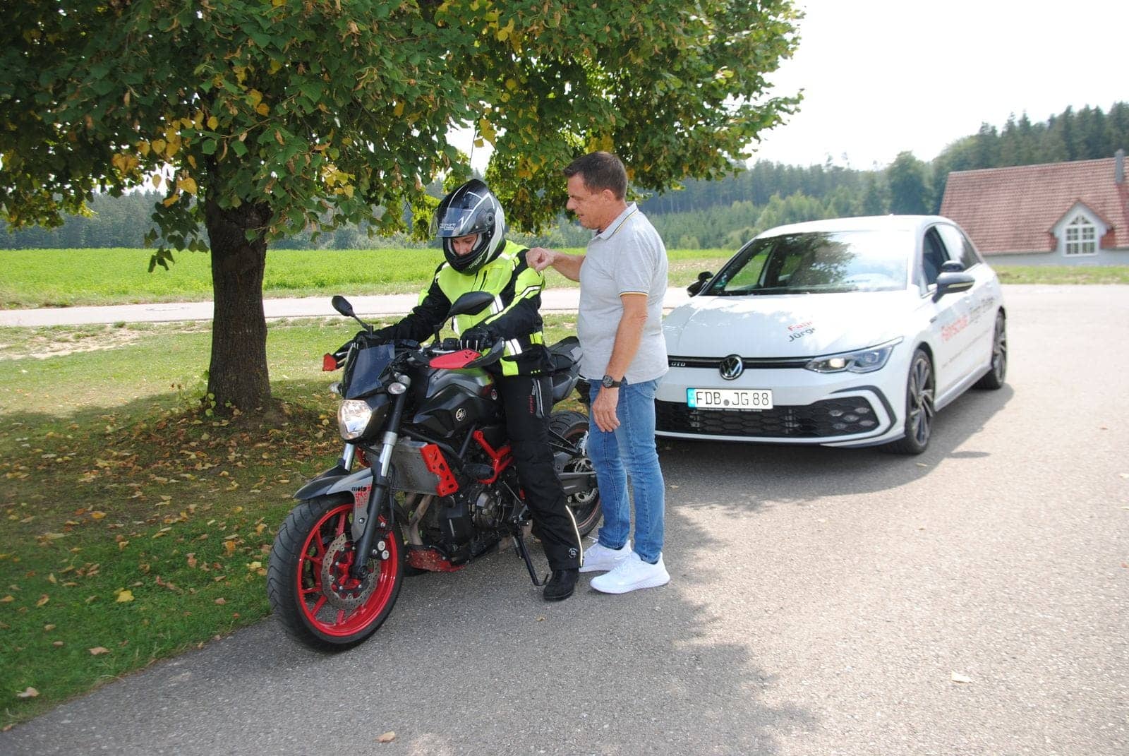 Fahrlehrer bespricht die Motorrad-Fahrstunde mit einer Fahrschülerin in Schutzkleidung, dahinter das Fahrschulauto